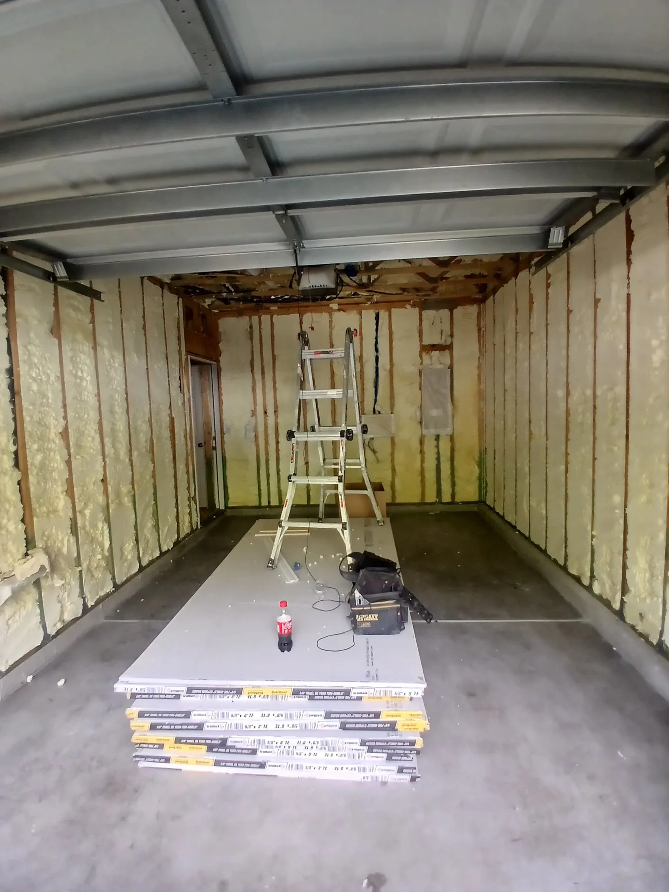 Garage interior with spray foam insulation, ladder, and drywall materials in progress